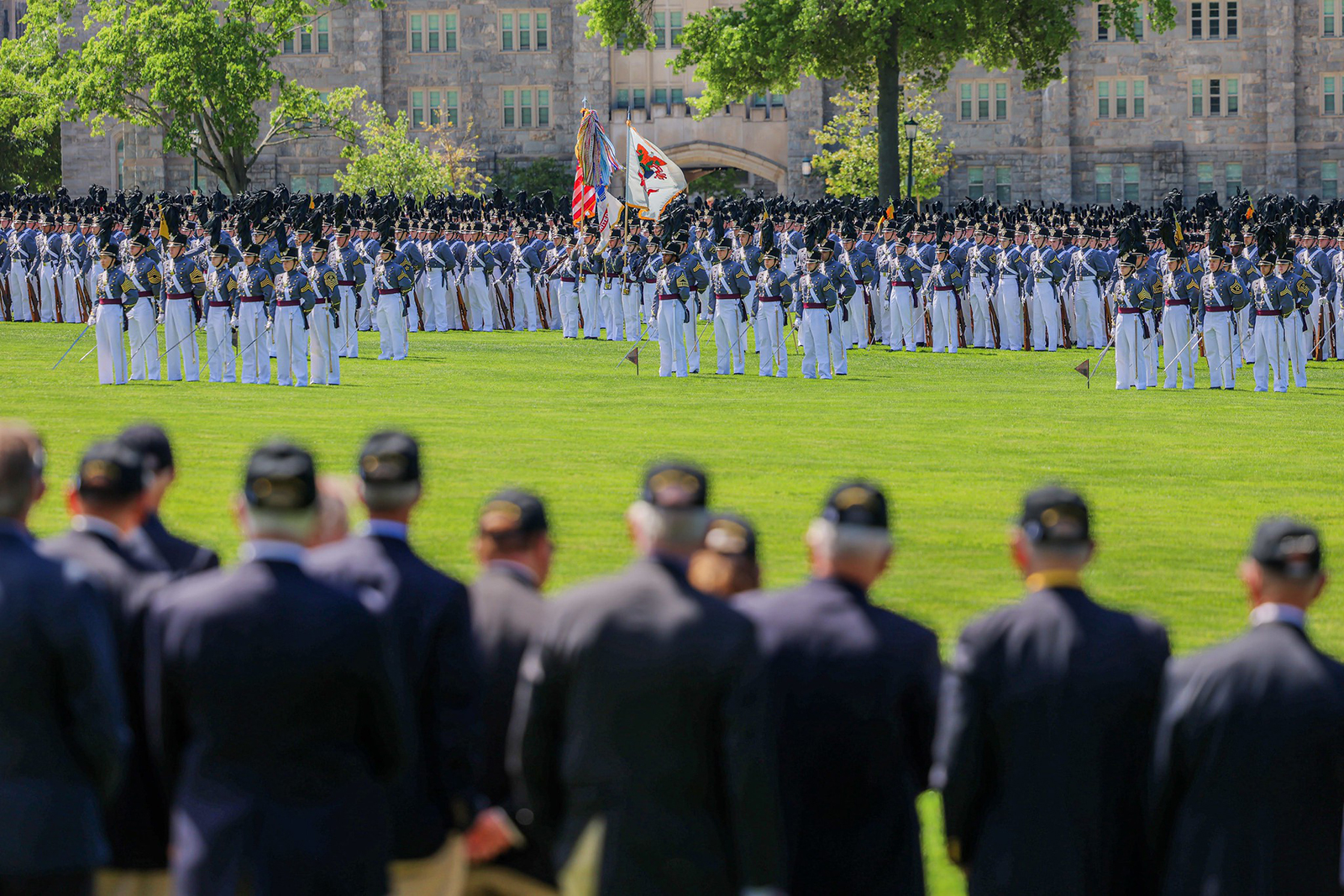 West Point cadets in full dress grey and white uniforms stand in formation on a parade field, viewed from behind a row of veterans watching the ceremony.